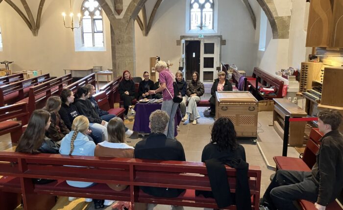 Schüler:innen sitzen in einer Kirche auf Kirchenbänken und bekommen die Funktion von Orgelpfeifen erklärt