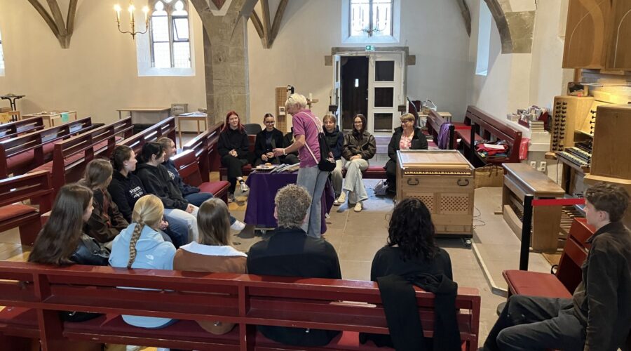 Schüler:innen sitzen in einer Kirche auf Kirchenbänken und bekommen die Funktion von Orgelpfeifen erklärt